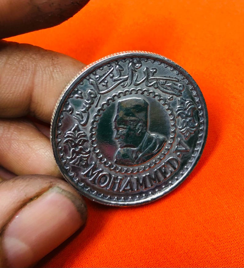 May include: A silver coin with a profile of a man wearing a hat and the word "MOHAMMED" inscribed around the edge. The coin also features Arabic script.