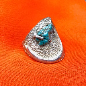 Peut inclure: Une bague en argent avec une surface texturée et une forme triangulaire. La bague présente une pierre turquoise au centre. La bague est posée sur un fond orange.
