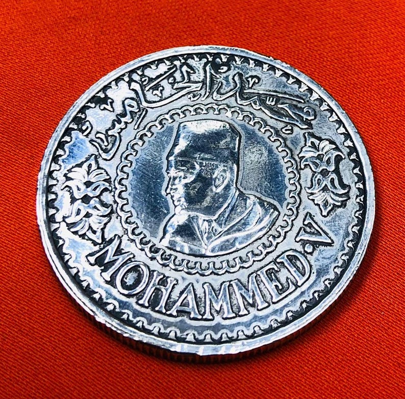 May include: Silver coin with a portrait of a man wearing a hat and the text "MOHAMMED V" around the edge.