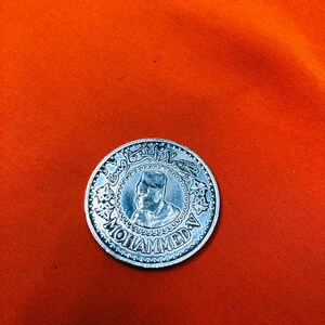 May include: A silver coin with a portrait of a man wearing a hat and the word "MOHAMMED" inscribed around the edge.