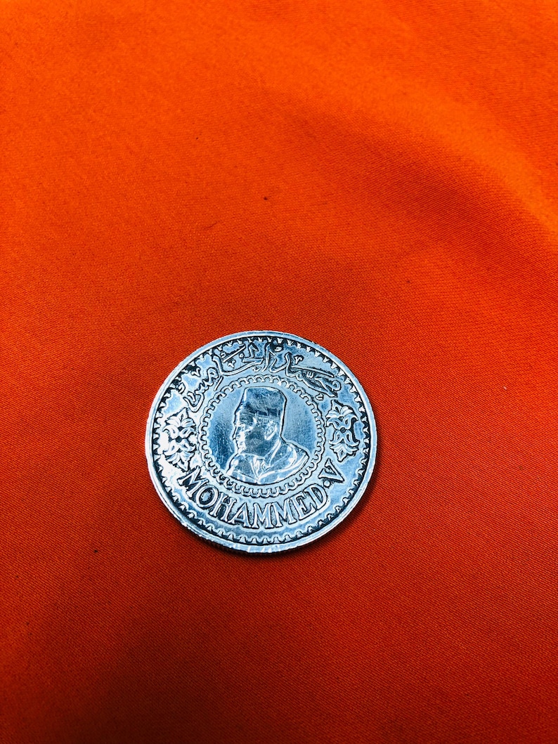 May include: A silver coin with a portrait of a man wearing a hat and the word "MOHAMMED" inscribed around the edge.