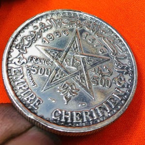May include: A silver coin with a five-pointed star in the center. The coin has Arabic text around the edge and the words "EMPIRE CHERIFIEN" and "500 FRANCS" on the bottom. The year "1956" is visible at the top of the coin.