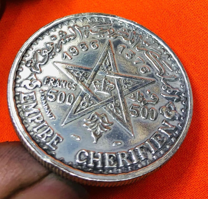 May include: A silver coin with a five-pointed star in the center. The coin has Arabic text around the edge and the words "EMPIRE CHERIFIEN" and "500 FRANCS" on the bottom. The year "1956" is visible at the top of the coin.