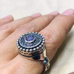 Puede incluir: Un anillo de plata con un diseño circular detallado. El anillo presenta un patrón floral central en azul, rojo y blanco, rodeado de intrincados trabajos en metal y pequeños detalles de cuentas. El anillo se lleva en un dedo.