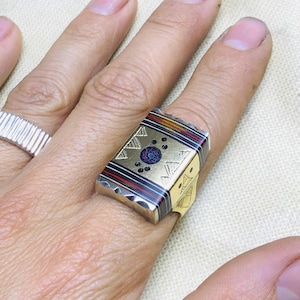 Puede incluir: Un anillo rectangular con detalles dorados, plateados, rojos y negros. El anillo presenta un diseño central con un patrón floral azul y rojo, rodeado de formas geométricas doradas y puntos negros. El anillo está en un dedo.