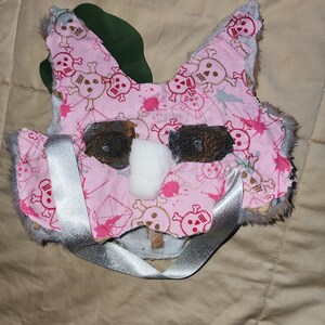 May include: Pink and white plush mask with a skull and crossbones pattern. The mask has a silver ribbon tied around it.