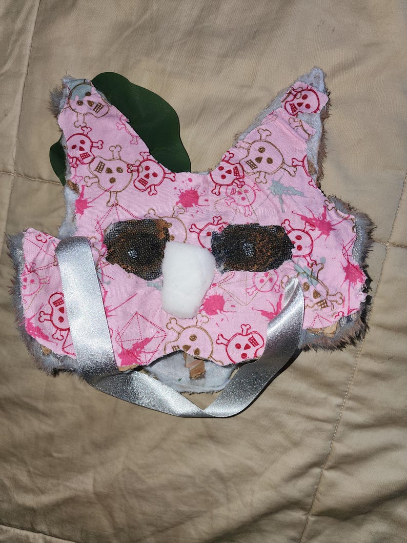 May include: Pink and white plush mask with a skull and crossbones pattern. The mask has a silver ribbon tied around it.