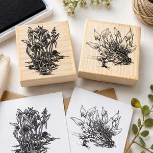 May include: Two square wooden rubber stamps featuring black ink illustrations of a duck in reeds and a frog among plants. Below the stamps are two white cards displaying the same black ink designs, showing the stamped images on paper.