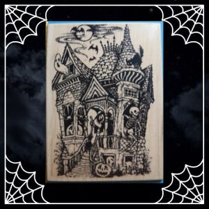 May include: A rectangular wooden rubber stamp featuring a detailed black ink illustration of a haunted house. The design includes a skeleton, a ghost, a witch, a black cat, bats, a full moon, and a jack-o'-lantern on the front porch steps.