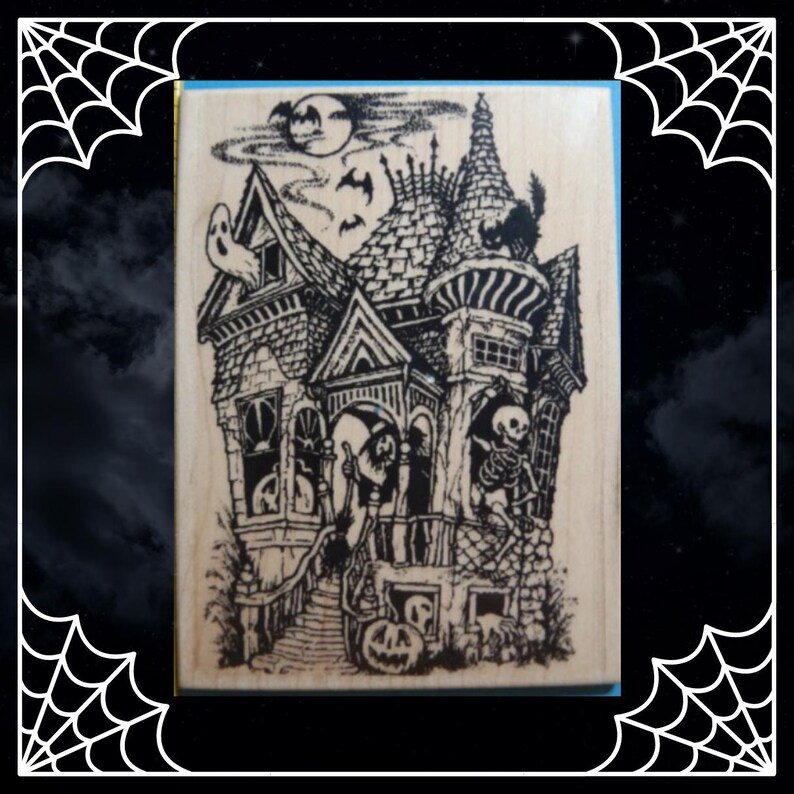 May include: A rectangular wooden rubber stamp featuring a detailed black ink illustration of a haunted house. The design includes a skeleton, a ghost, a witch, a black cat, bats, a full moon, and a jack-o'-lantern on the front porch steps.