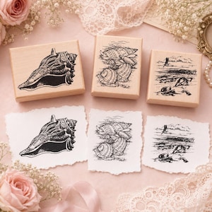 May include: Three rectangular wooden rubber stamps and three corresponding paper prints. The stamps feature black ink illustrations of a large conch shell, a cluster of various sea shells, and a coastal beach scene with a bird flying overhead.