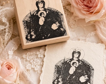 May include: A square wooden rubber stamp featuring a black ink illustration of a crowned figure holding a child, seated on a throne. Beside the stamp is a square piece of textured off-white paper displaying the same black ink stamped image.