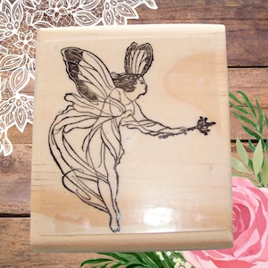 May include: A wooden stamp featuring a detailed black illustration of a fairy with butterfly wings, holding a wand. The fairy is depicted in a flowing gown. The stamp is rectangular with a clear, transparent base.