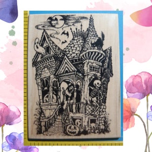 May include: A rectangular wooden rubber stamp featuring a detailed black ink illustration of a haunted house with a ghost, a skeleton, a black cat, and a jack-o'-lantern. A yellow measuring tape is visible along the left edge of the stamp.