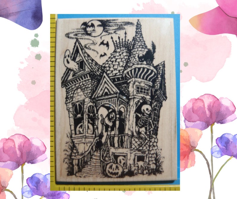 May include: A rectangular wooden rubber stamp featuring a detailed black ink illustration of a haunted house with a ghost, a skeleton, a black cat, and a jack-o'-lantern. A yellow measuring tape is visible along the left edge of the stamp.