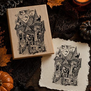 May include: A rectangular wooden rubber stamp featuring a detailed black ink illustration of a haunted Victorian house with a skeleton, a ghost, and a jack-o'-lantern, accompanied by a matching stamped image on a piece of textured off-white paper.