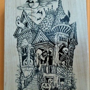 May include: A rectangular wooden block featuring a black ink illustration of a haunted house. The scene includes a ghost in a window, a witch on the porch, a skeleton, a black cat on the roof, bats, a full moon, and a jack-o'-lantern.
