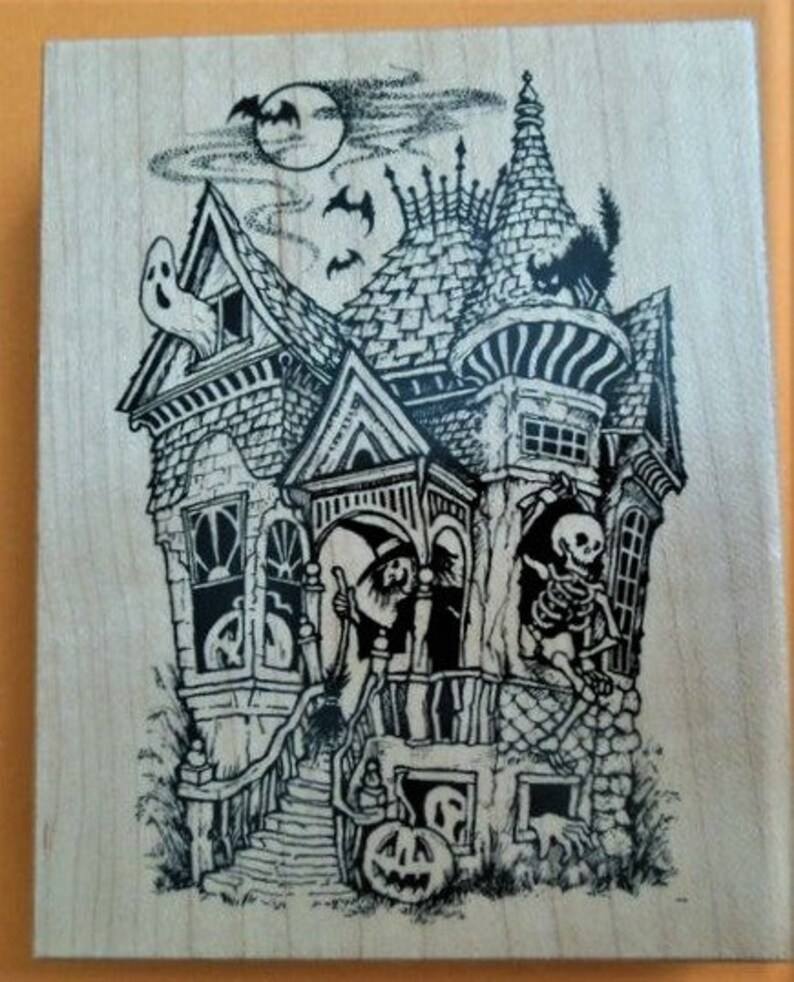 May include: A rectangular wooden block featuring a black ink illustration of a haunted house. The scene includes a ghost in a window, a witch on the porch, a skeleton, a black cat on the roof, bats, a full moon, and a jack-o'-lantern.
