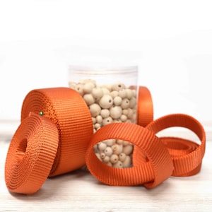 May include: Orange webbing or strapping in various sizes and shapes. The webbing is rolled up and some pieces are loose. A clear jar filled with white wooden beads is in the background.