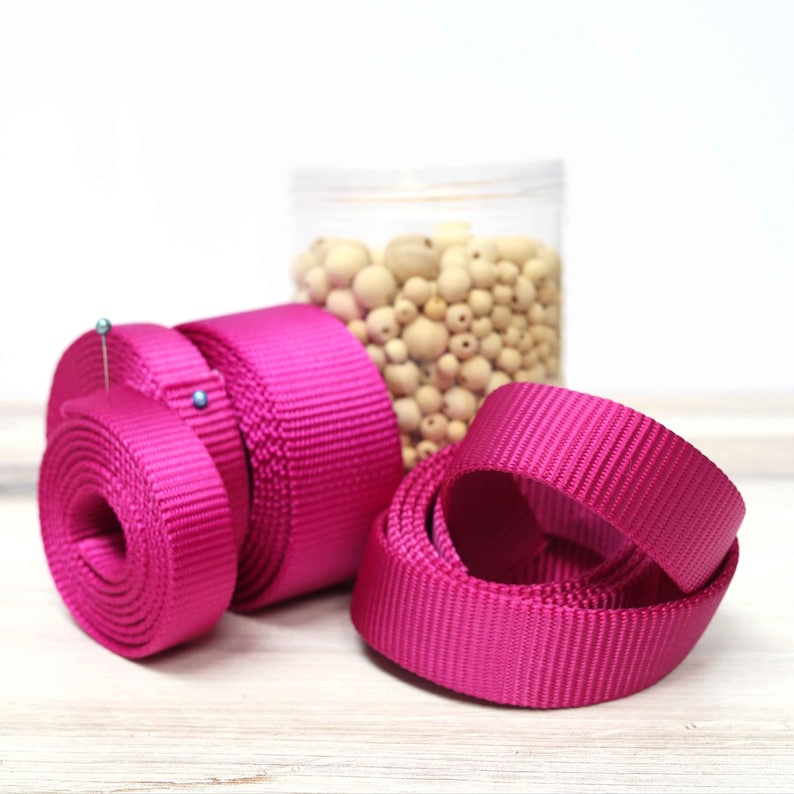 May include: Three spools of bright pink nylon webbing, each with a different width. The spools are arranged on a white surface.