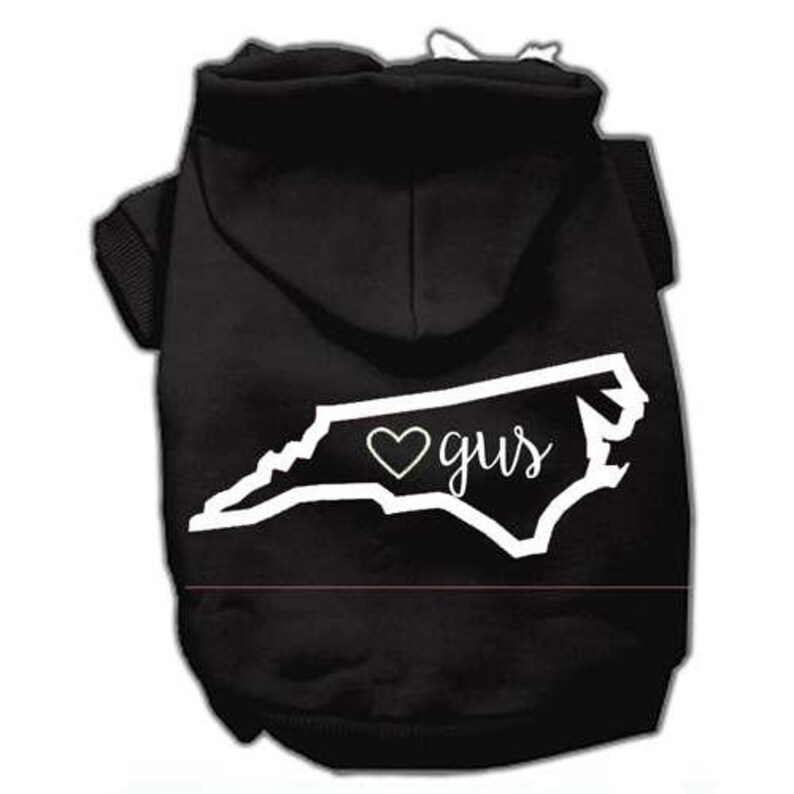 Puede incluir: Sudadera con capucha negra para perro con un contorno blanco del estado de Carolina del Norte y un coraz&oacute;n que contiene el nombre "Gus".