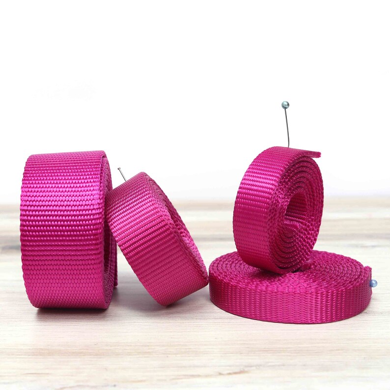 May include: Three rolls of pink nylon webbing, each with a different width. The webbing is rolled up and the ends are secured with a pin.