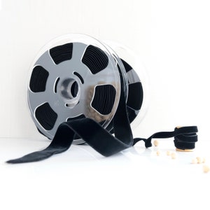 May include: A spool of black velvet ribbon wound around a clear plastic spool. The ribbon is lying on a white surface.