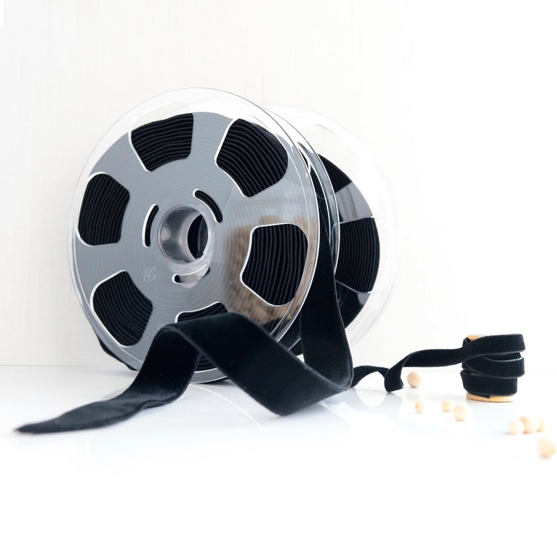 May include: A spool of black velvet ribbon wound around a clear plastic spool. The ribbon is lying on a white surface.