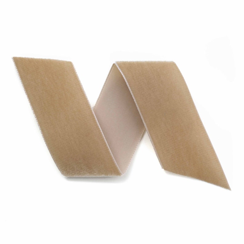 Puede incluir: Una cinta de terciopelo color beige, doblada en zigzag sobre un fondo blanco. La cinta tiene una textura suave y afelpada, con un reverso de color m&aacute;s claro. Ideal para manualidades, costura o envolver regalos.
