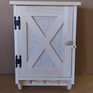 May include: A small, unfinished wooden cabinet with a single door and a decorative X-shaped design. The cabinet has a shelf inside and hooks on the bottom.