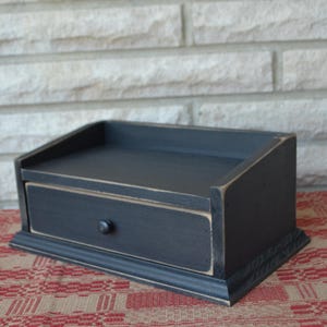 May include: A small, black wooden desk organizer with a single drawer and a sloped top. The organizer has a distressed finish, giving it a rustic appearance. The drawer features a round, dark knob. The piece sits on a decorative base.