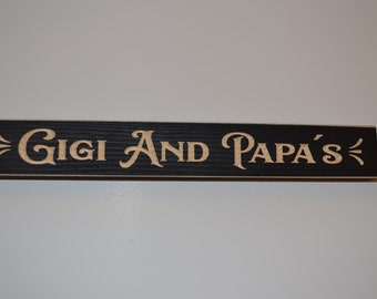 May include: A black wooden sign with the text "Gigi and Papa's" in a gold-colored font.
