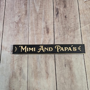 May include: Black wooden sign with gold lettering that reads "Mimi and Papa's".