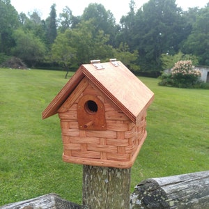 May include: A wooden birdhouse with a woven design, painted in a reddish-brown color, mounted on a wooden fence post.