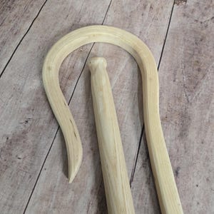 May include: A wooden shepherd's crook with a curved handle and a straight shaft. The crook is made of light brown wood and has a smooth finish.
