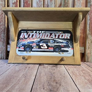 May include: Wooden shelf with a framed metal sign featuring a race car with the number 3. The sign reads "The Intimidator" and "Goodwrench Service Plus". The shelf has two small wooden pegs below the sign.