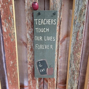 May include: A rustic wooden sign with a green background and white text that reads "Teachers Touch Our Lives Forever". The sign also features a small chalkboard with the words "1 + 1 = CAT" and a red apple.