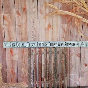 May include: A wooden sign with the text "I can do all things through Christ who strengthens me" painted in black. The sign is hanging on a wooden wall with a hay bale in the foreground.