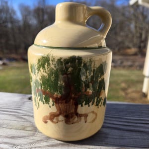 May include: A small, cream-colored ceramic jug with a green and brown tree design. The jug has a handle and a wide mouth.