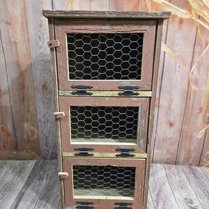 May include: A three-tiered wooden cabinet with wire mesh doors. The cabinet is painted in shades of brown and red, with black metal hinges. The cabinet is made of reclaimed wood and has a rustic look.