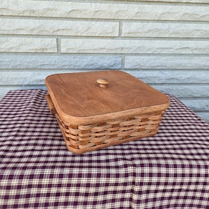 May include: A square woven basket with a wooden lid and a knob on top. The basket is brown and has a natural woven pattern. The basket is sitting on a red and white checkered tablecloth.
