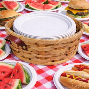 May include: A picnic scene with a red and white checkered tablecloth. The table is laden with plates of watermelon slices, hamburgers, hot dogs, corn on the cob, and potato salad. A woven basket holds a stack of white plates.