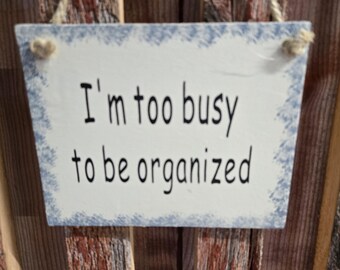 May include: A white wooden sign with blue edges hangs on a wooden fence. The sign reads "I'm too busy to be organized".