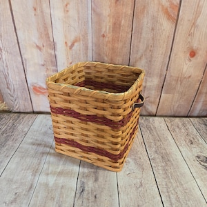 May include: A square woven basket with a brown and red striped pattern and a leather handle. The basket is sitting on a wooden surface.