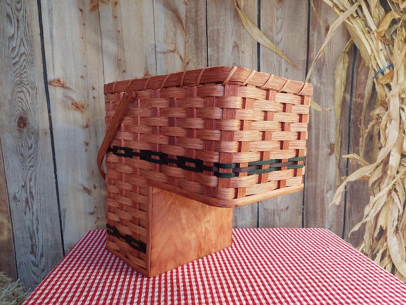 May include: A woven basket with a natural wood finish and black trim. The basket has a handle and is sitting on a red and white checkered tablecloth.