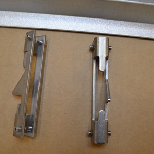 May include: Two silver metal brackets with a small opening in the center. The brackets have a small, angled piece of metal on the bottom.