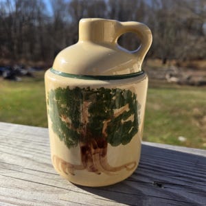 May include: A small, cream-colored ceramic pitcher with a green rim and a hand-painted design of a brown tree with green leaves.