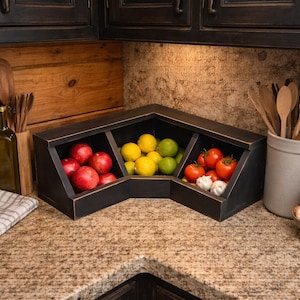 May include: A black wooden corner fruit and vegetable storage bin filled with red apples, yellow lemons, green limes, red tomatoes, and garlic. The bin is on a granite countertop in a kitchen setting.
