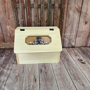 May include: A wooden bread box with a hinged lid and a clear window on the front. The window shows a painted design of a woman in a dress and a hat.