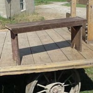 May include: A rustic, wooden bench with a dark brown finish. The bench has a rectangular seat and angled legs. The bench is approximately 48 inches long. The bench is outdoors, resting on a wooden platform.